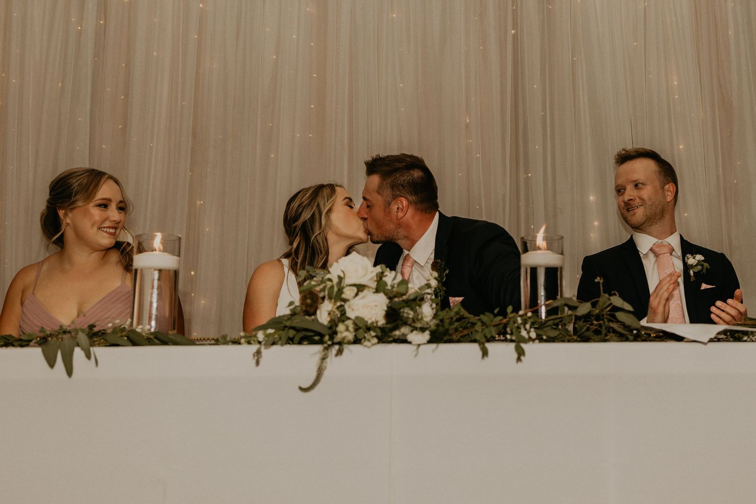 Wedding reception head table decorated with greenery and glowing candles.
