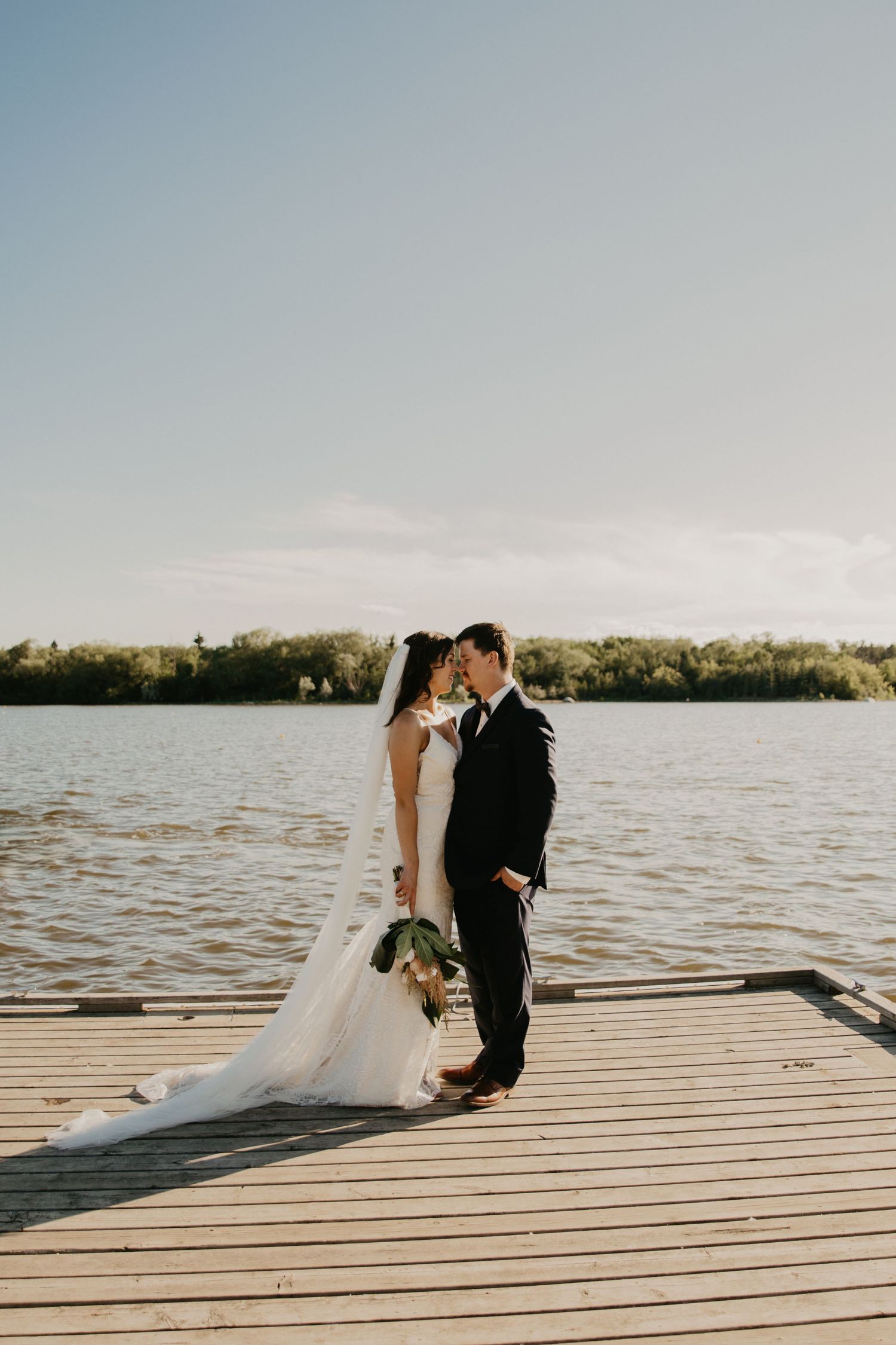 A romantic sunset wedding photo of a couple on a wooden dock by a serene lake.