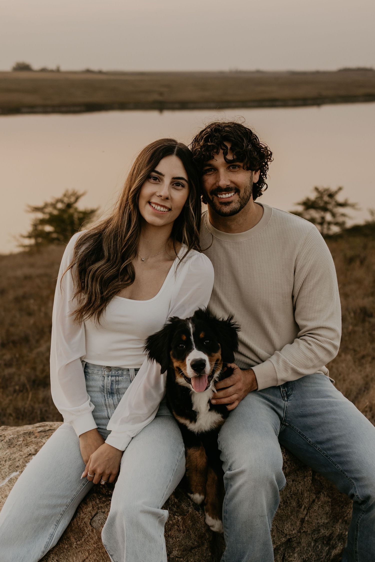 A couple sits on a rock with their Australian Shepherd dog against a sunset lake backdrop.