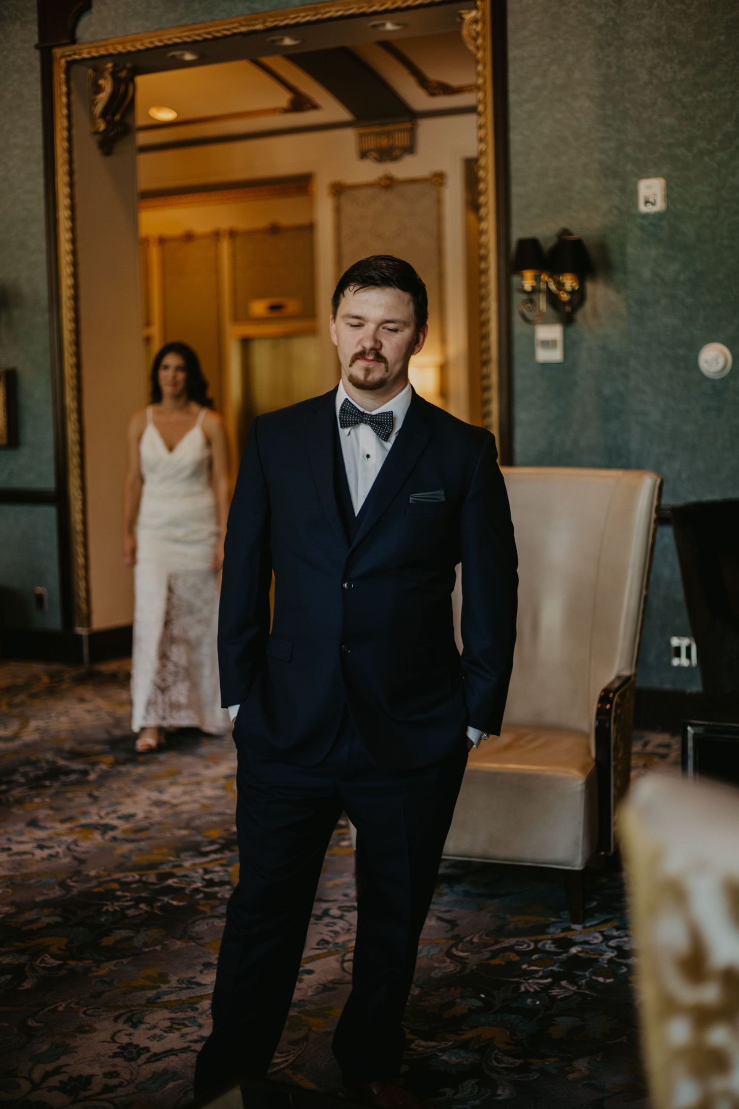 Wedding day scene showing a groom in formal black tuxedo with distant silhouette of bride in white dress.