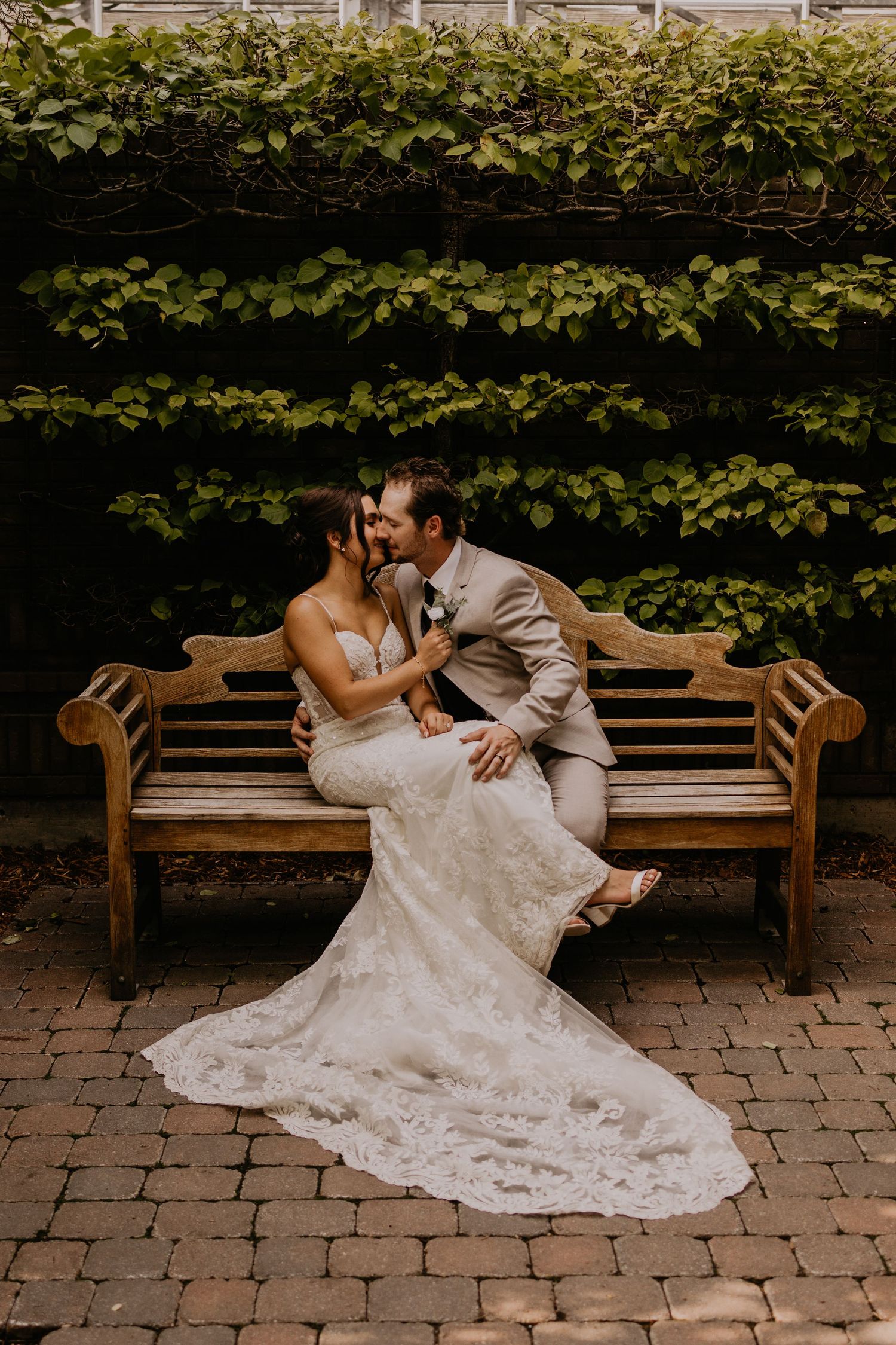 A couple in wedding attire sharing an intimate moment on a wooden bench against an ivy-covered wall.