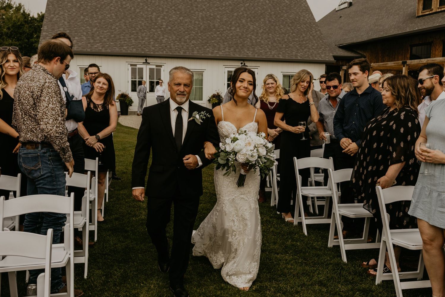 A wedding ceremony processional takes place on a lawn with white chairs and guests watching.