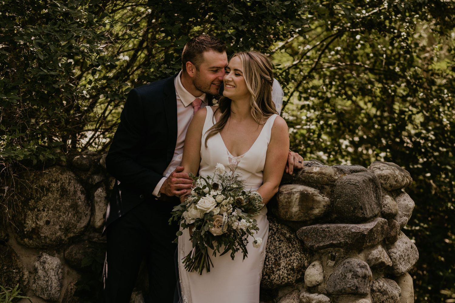 Romantic moment captured against rustic stone wall with elegant white bouquet.
