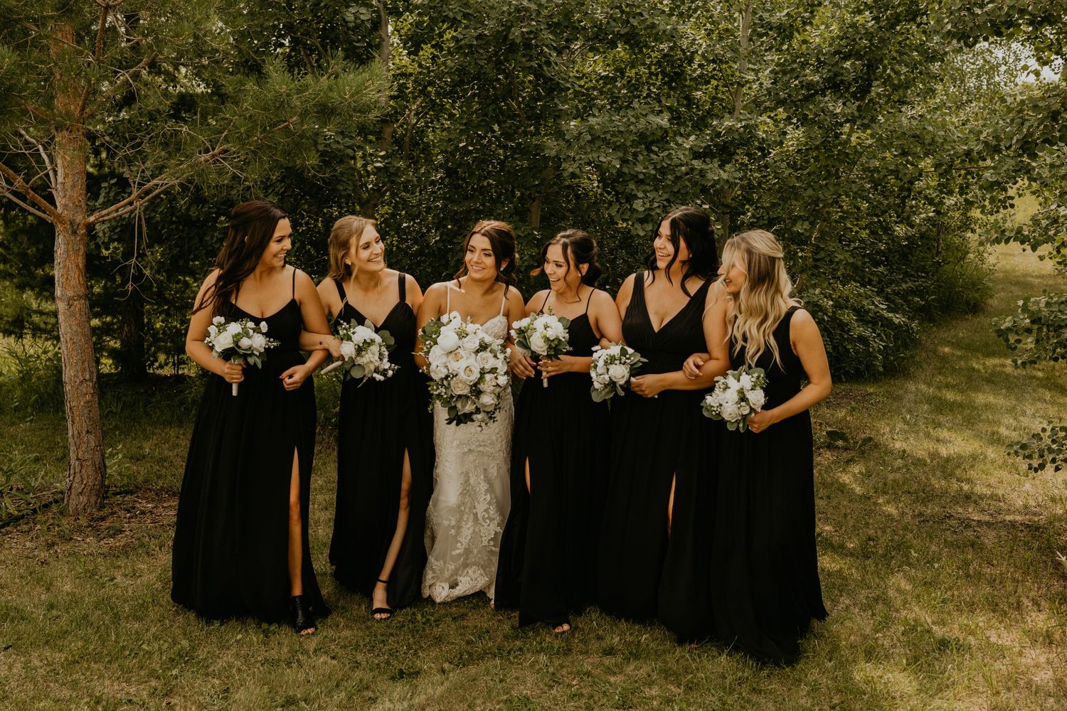 Group photo of bridal party in black dresses and white lace gown gathered on grass with trees behind.