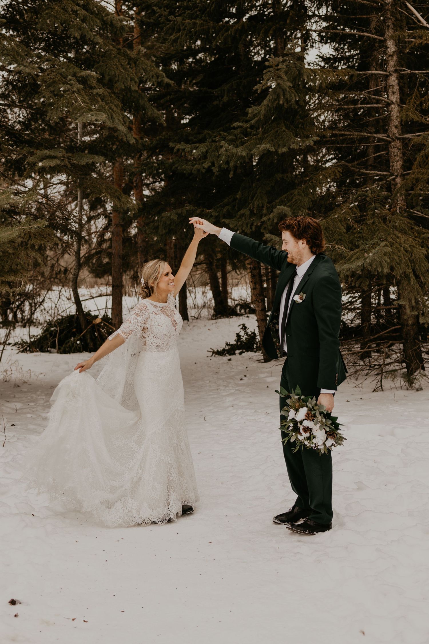 A couple share a playful moment in a snowy forest setting with the bride twirling in her lace wedding gown.
