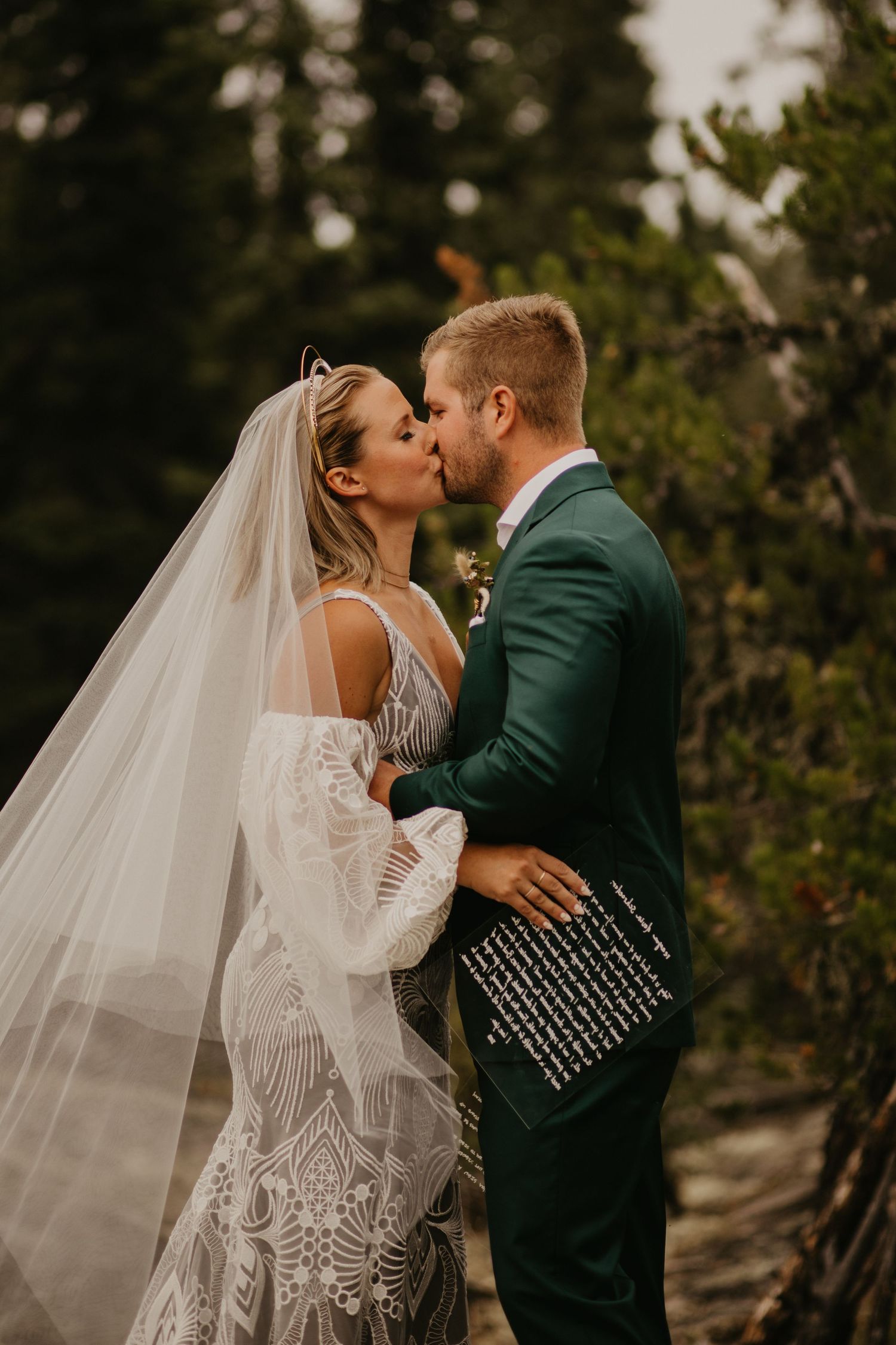 Romantic wedding kiss captured in golden light with forest backdrop.