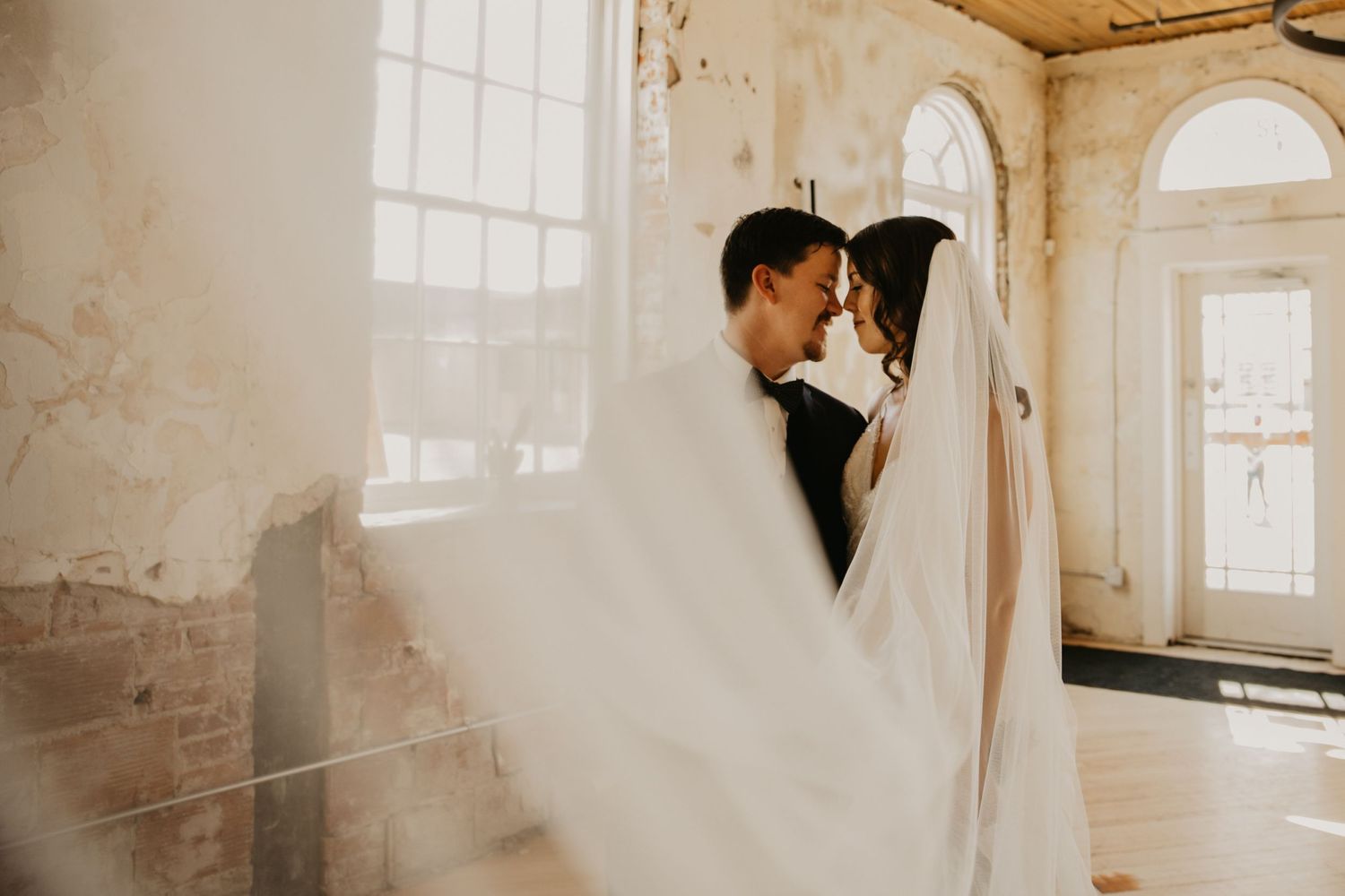 An ethereal wedding photo with flowing veil in a rustic room with arched windows.