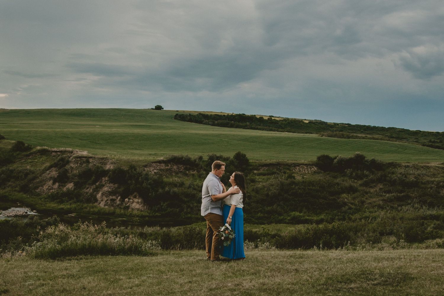 A romantic couple embraces on a grassy hilltop against a moody sky.