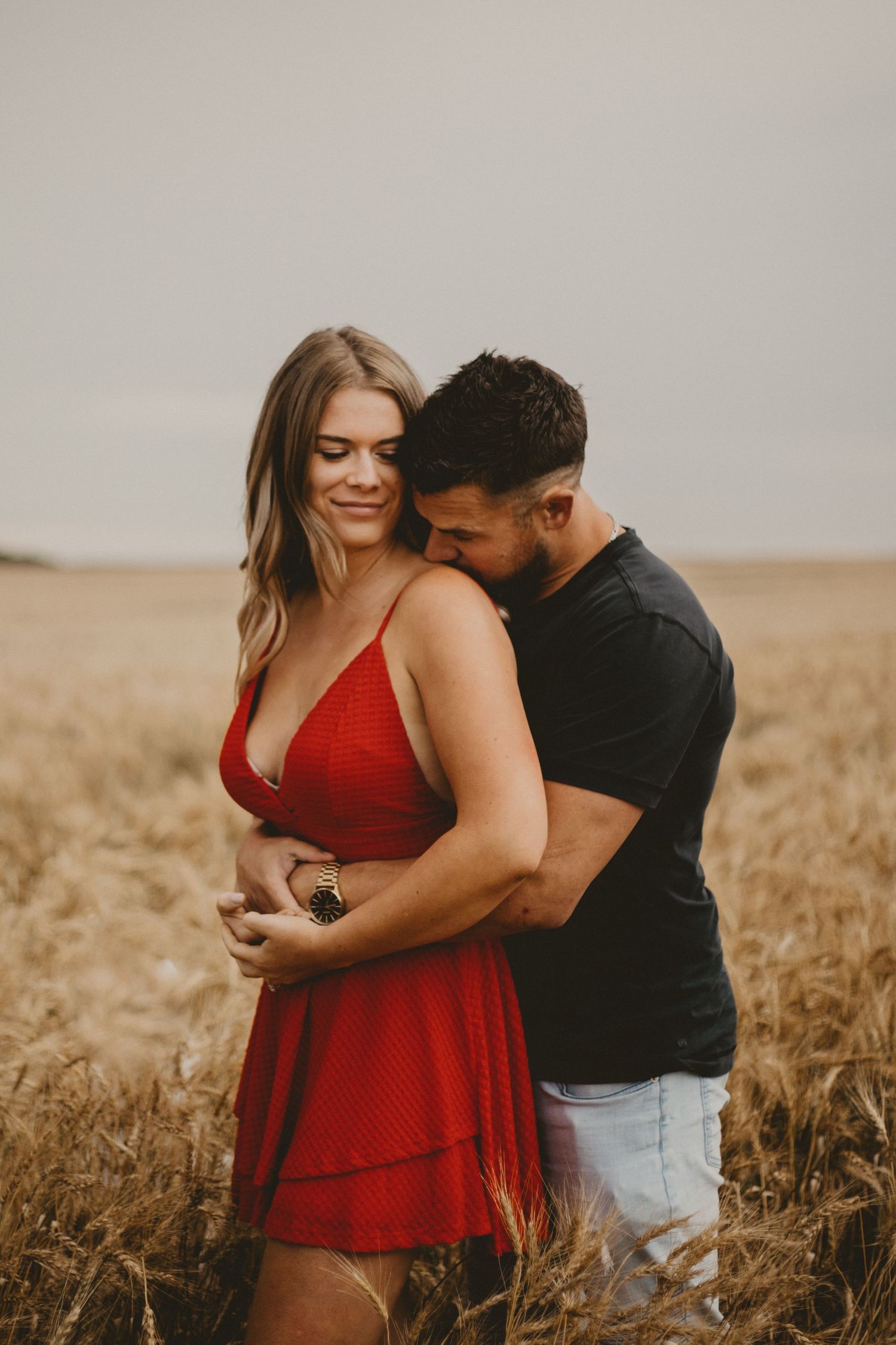 Couple embraces intimately in a golden wheat field at sunset with the woman wearing a red dress.
