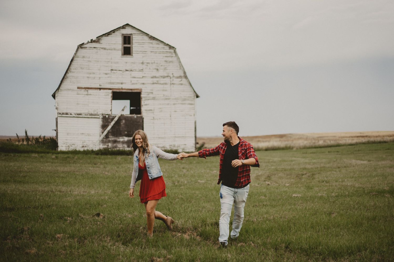 A couple playfully runs in front of a rustic white barn in a rural setting.