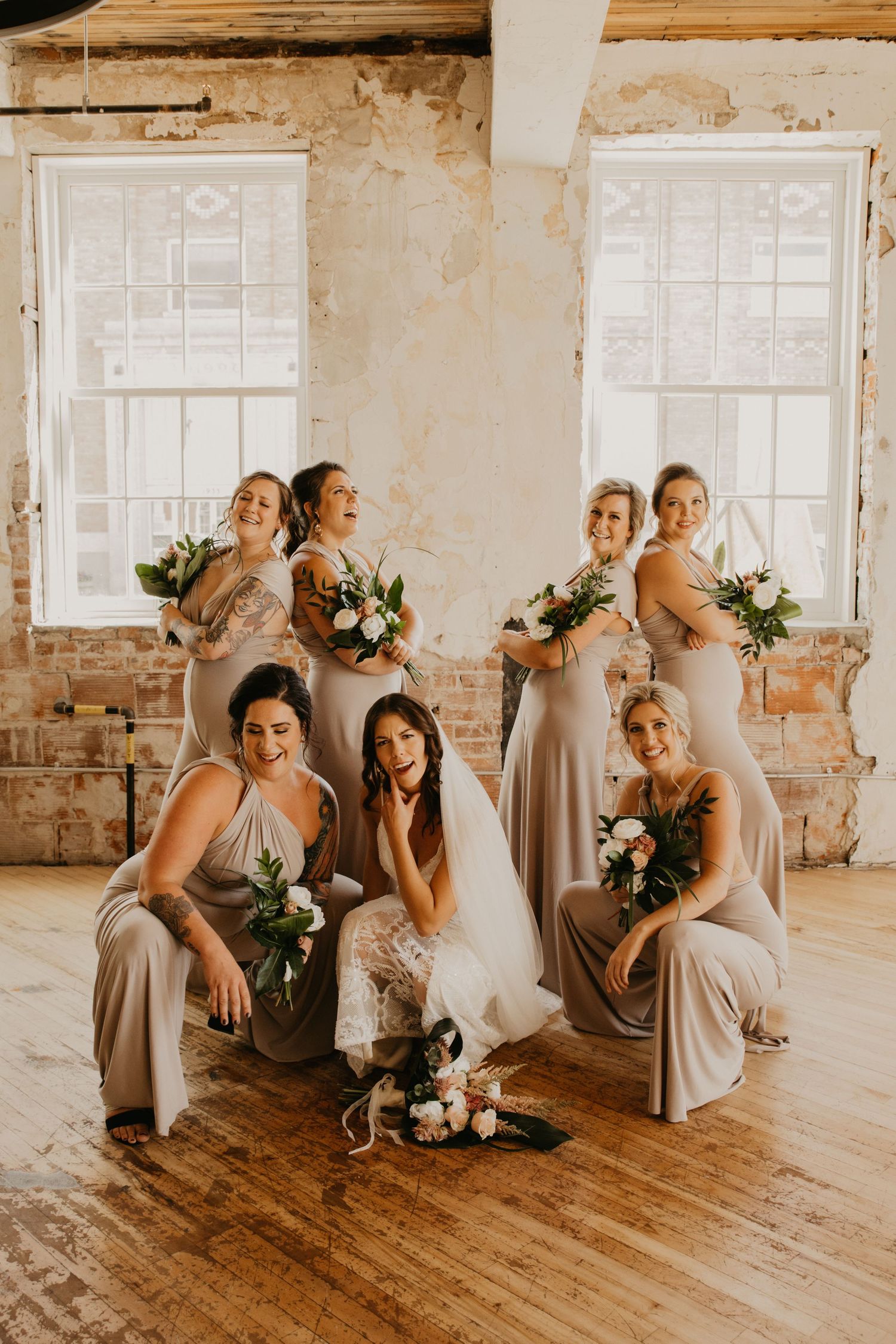 Wedding party in beige dresses poses together with bouquets in an industrial-style room.
