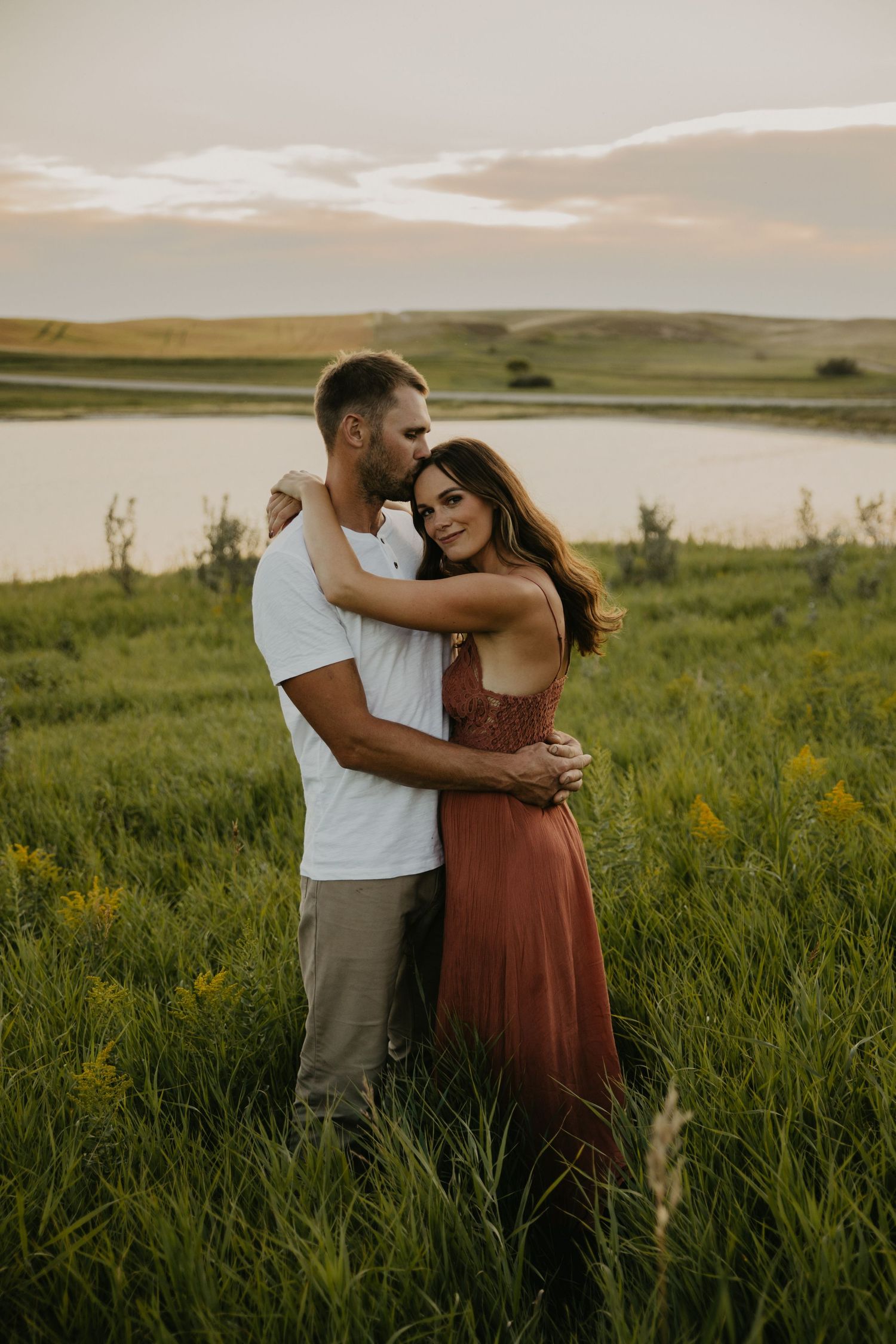 A romantic couple embraces in tall grass with a serene lake backdrop at sunset.