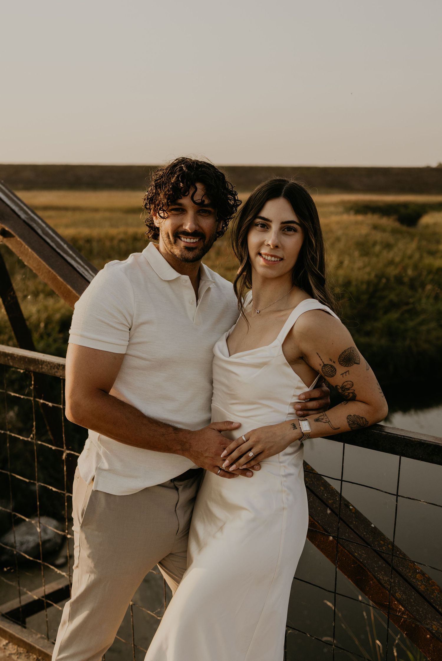 A romantic couple poses together on a scenic overlook during golden hour in casual elegant attire.