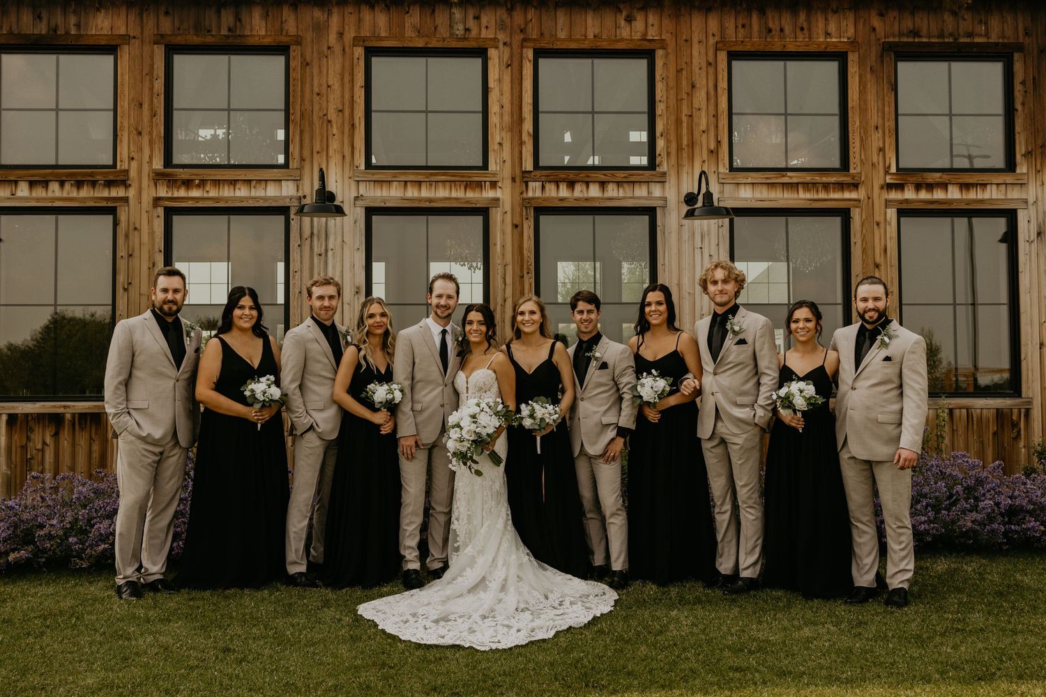 Wedding party in coordinated attire celebrates outside a rustic venue with wooden paneling.