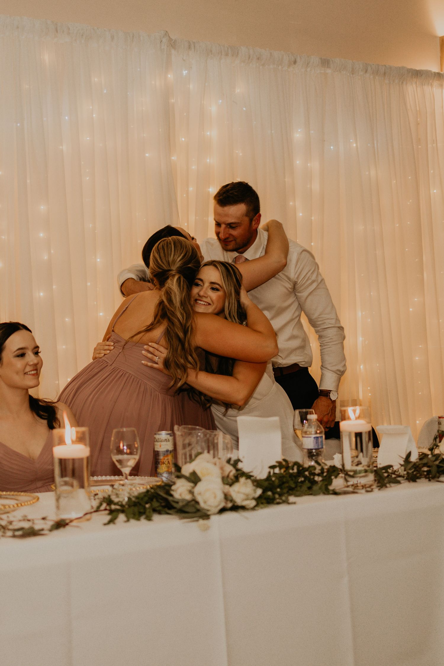 Wedding reception guests embrace at a decorated head table with white linens and floral centerpieces.