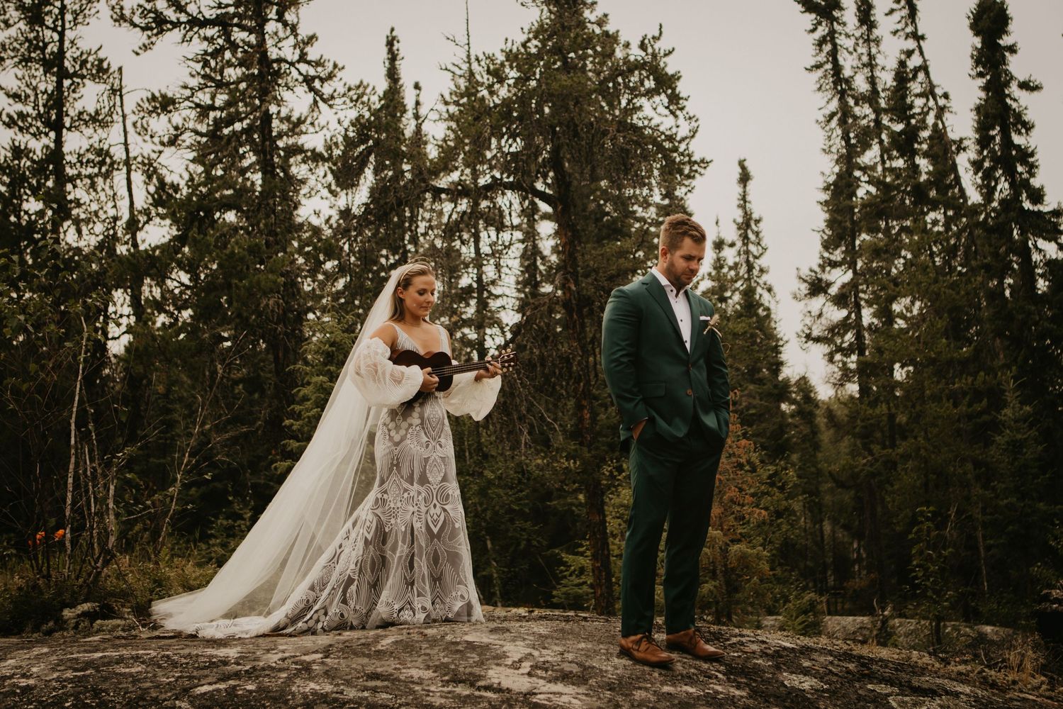 A bride and groom pose separately on a rocky outcrop surrounded by towering pine trees.
