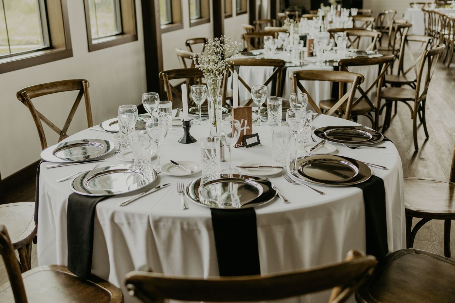 Wedding reception table setup with white linens, cross-back chairs, and elegant place settings.
