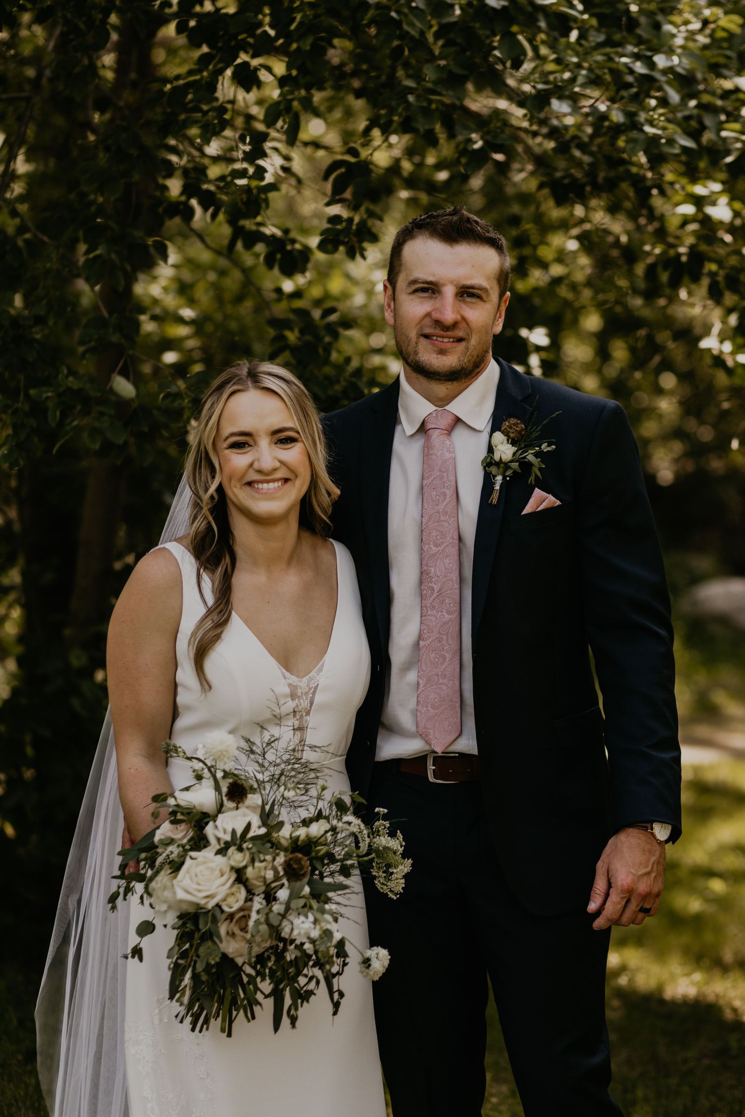A wedding couple poses outdoors under trees with the bride holding a lush white and greenery bouquet.