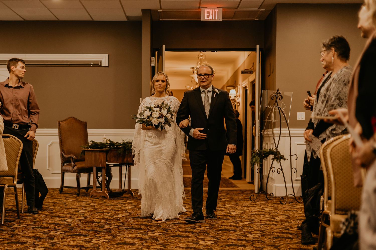A couple make their entrance into a carpeted ballroom during a wedding ceremony with warm lighting and guests watching.