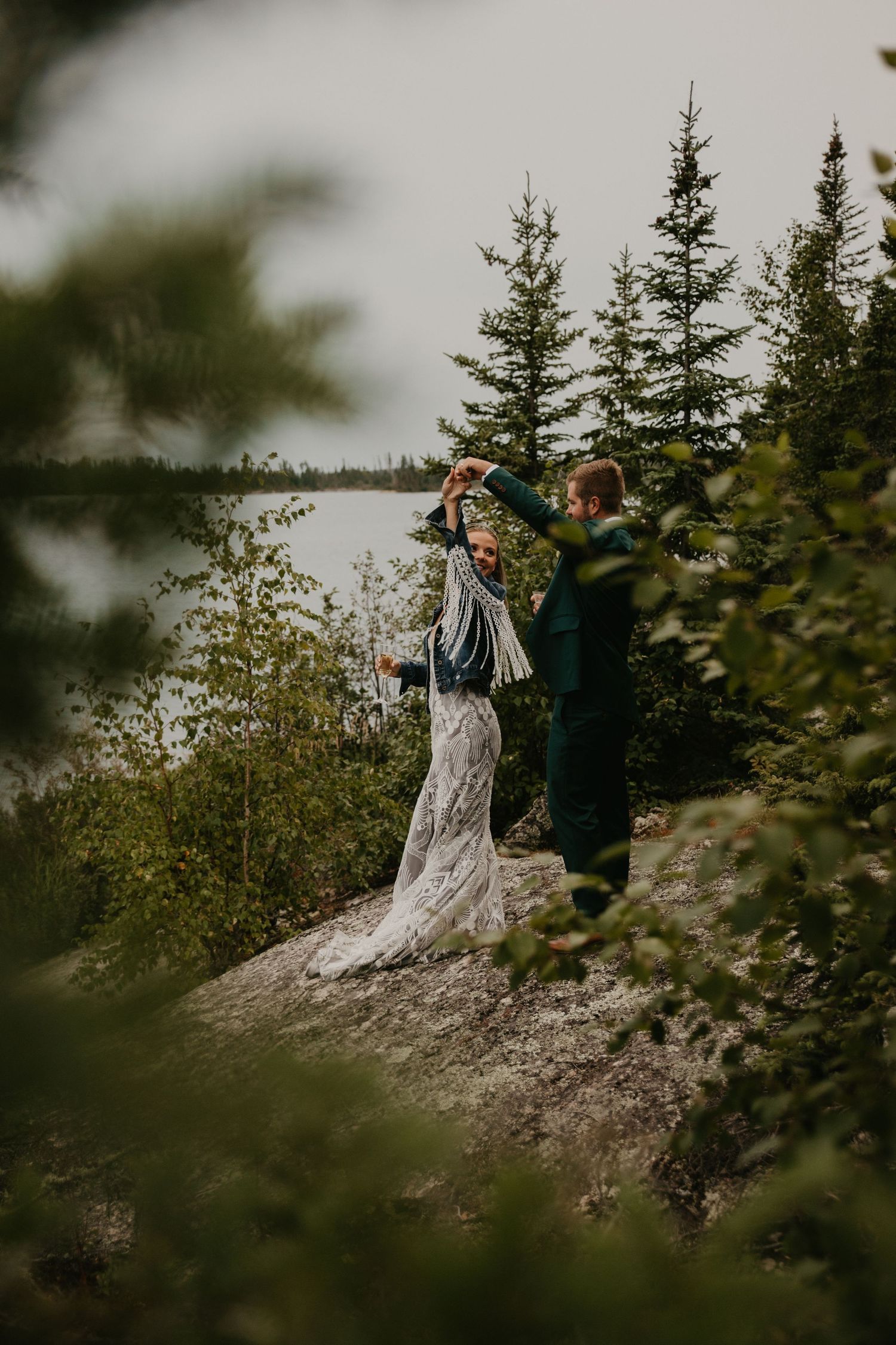 A couple stands on a cliff edge overlooking a lake surrounded by evergreen forest.
