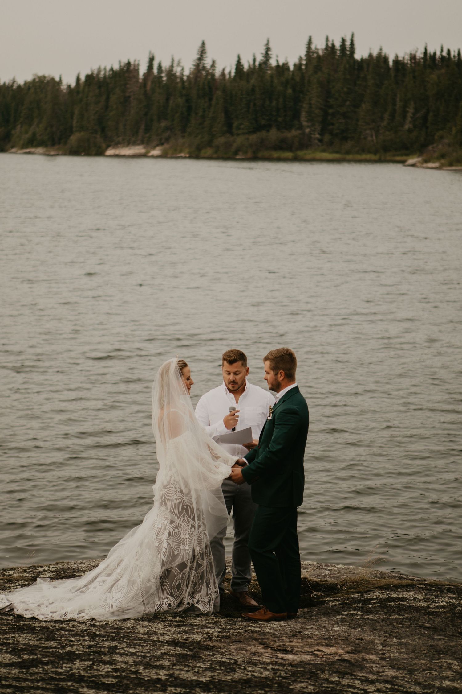A lakeside wedding ceremony taking place on rocky shore with evergreen trees in the background.