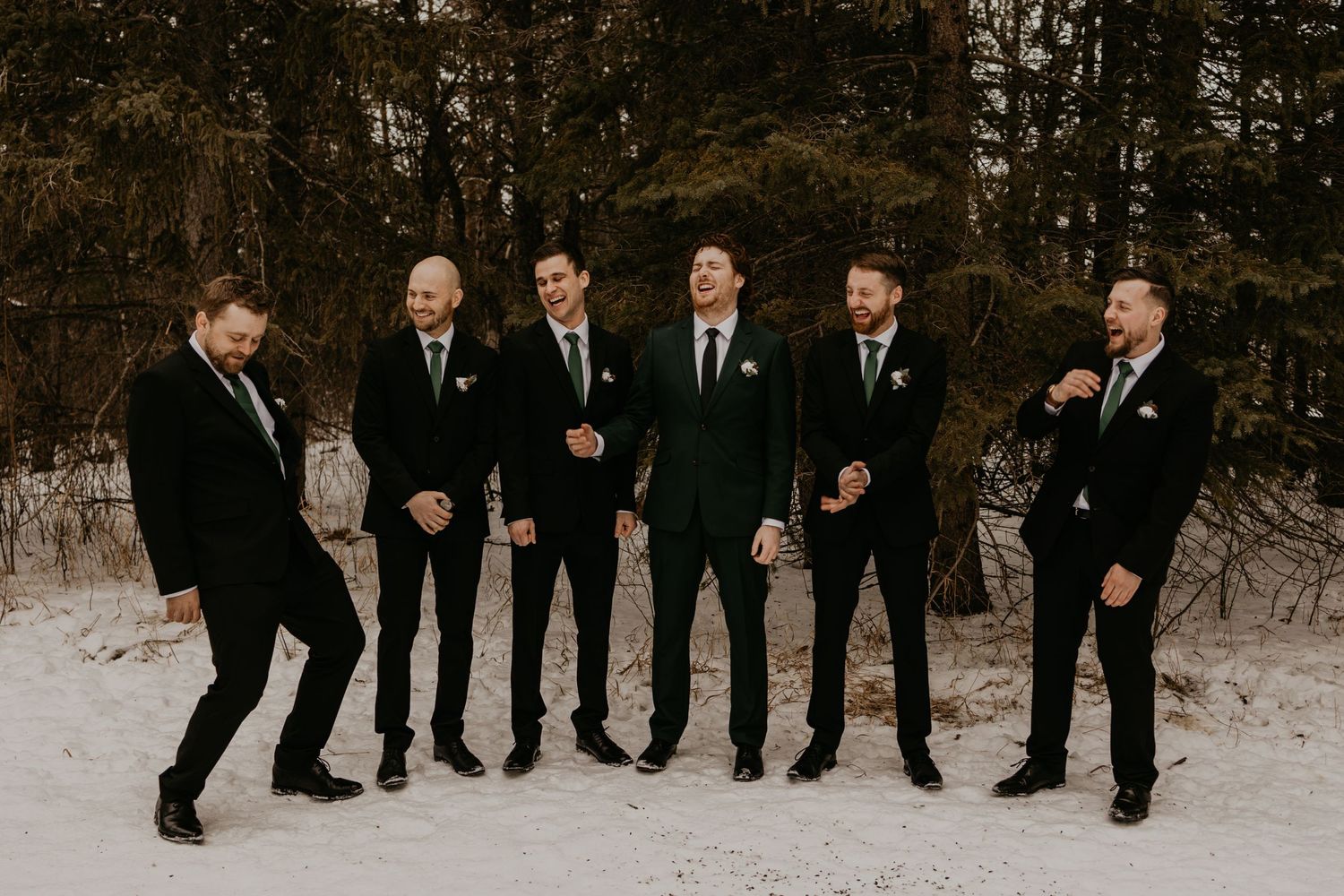 Groomsmen in black suits pose casually on snow-covered ground with evergreen forest backdrop.