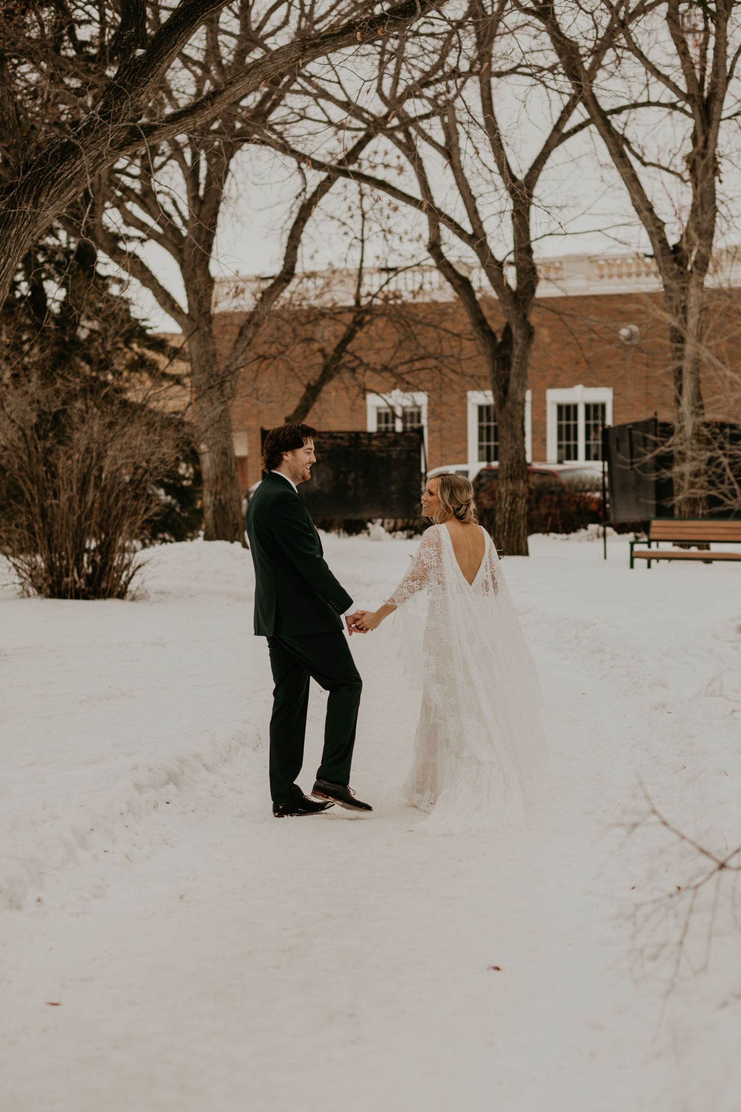 A winter wedding scene with a couple dancing in the snow near a brick building.