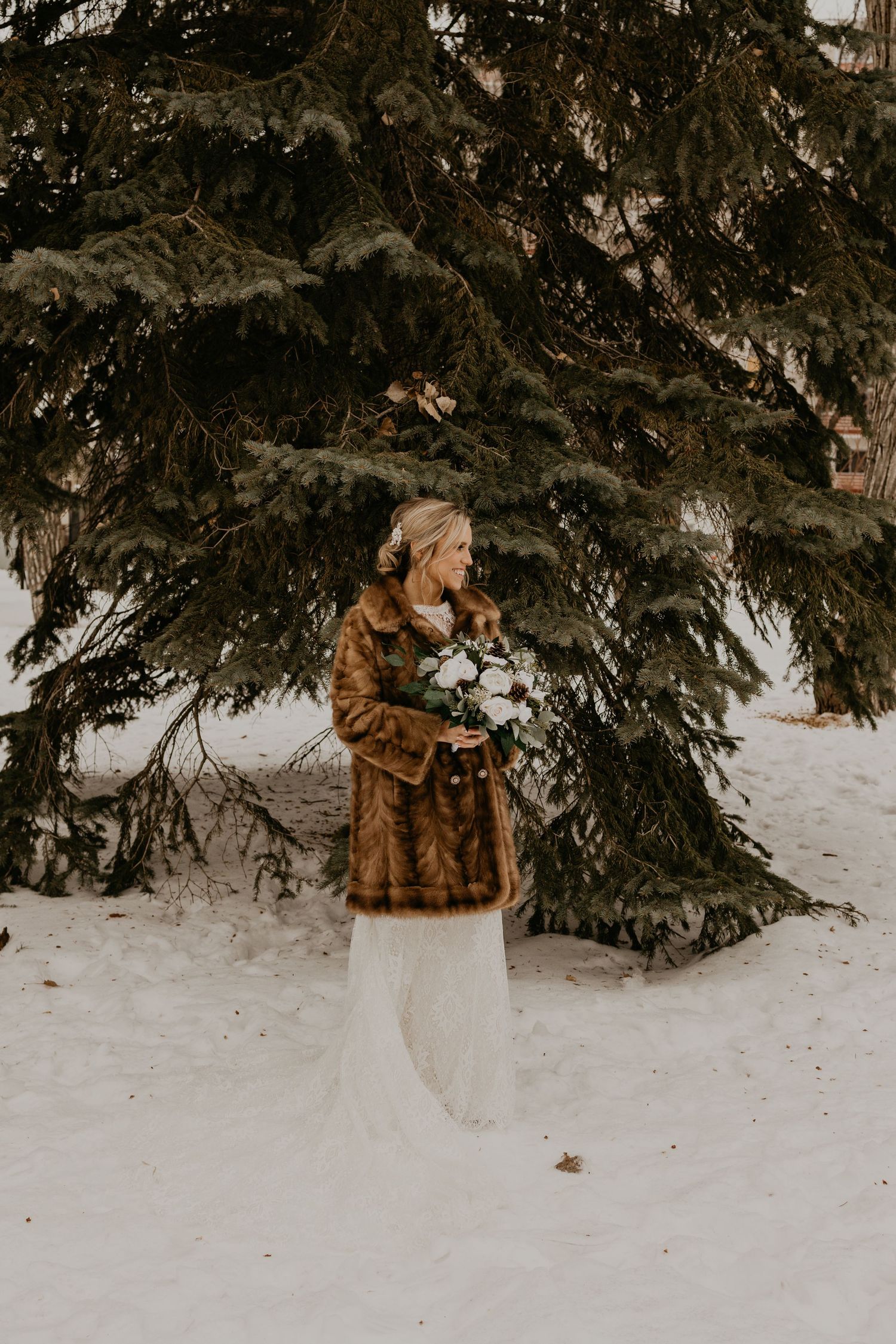 Bride in a white dress and brown fur coat holds a bouquet while standing in snow next to evergreen trees.