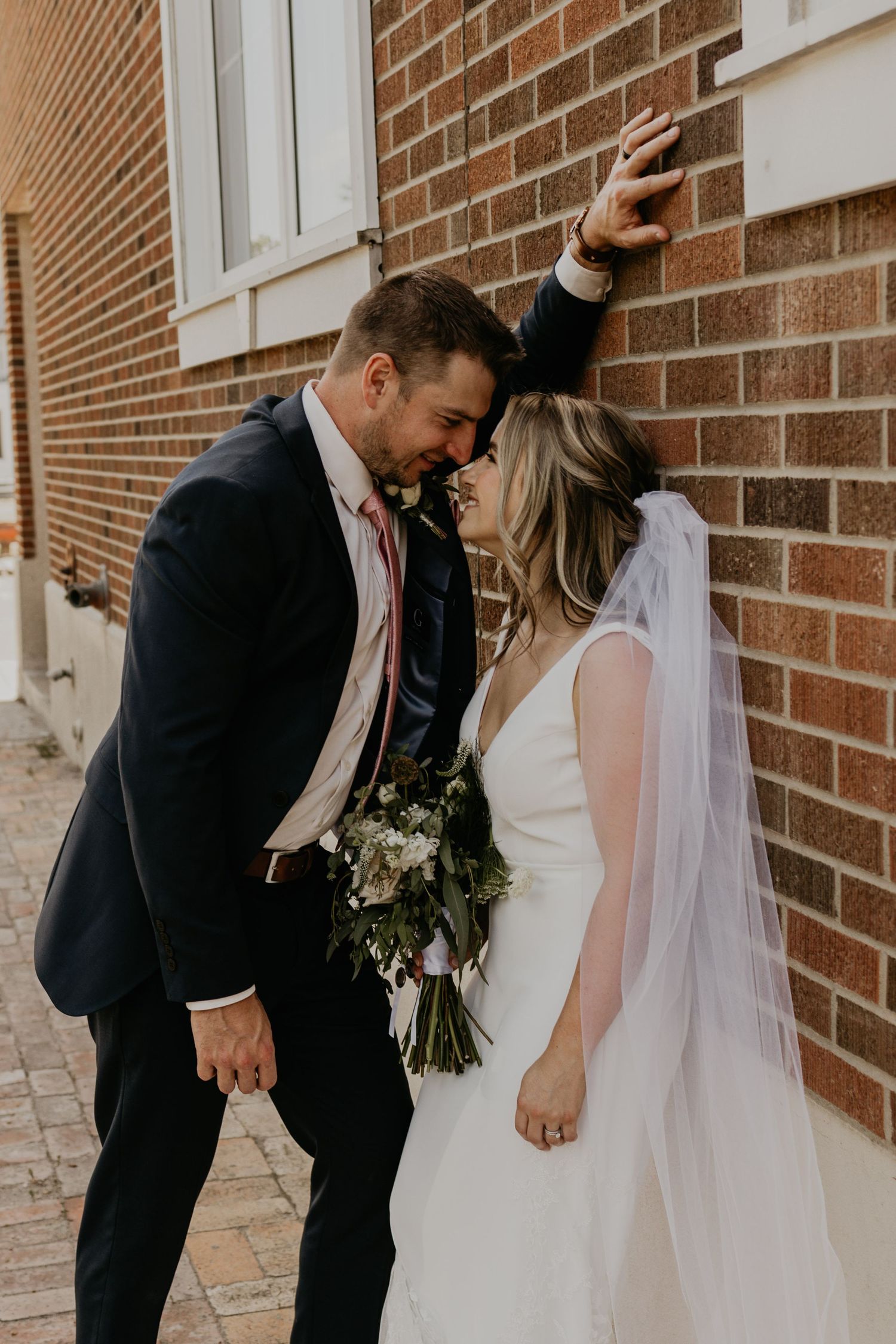 A romantic wedding moment captured against a brick building with a bouquet of flowers.