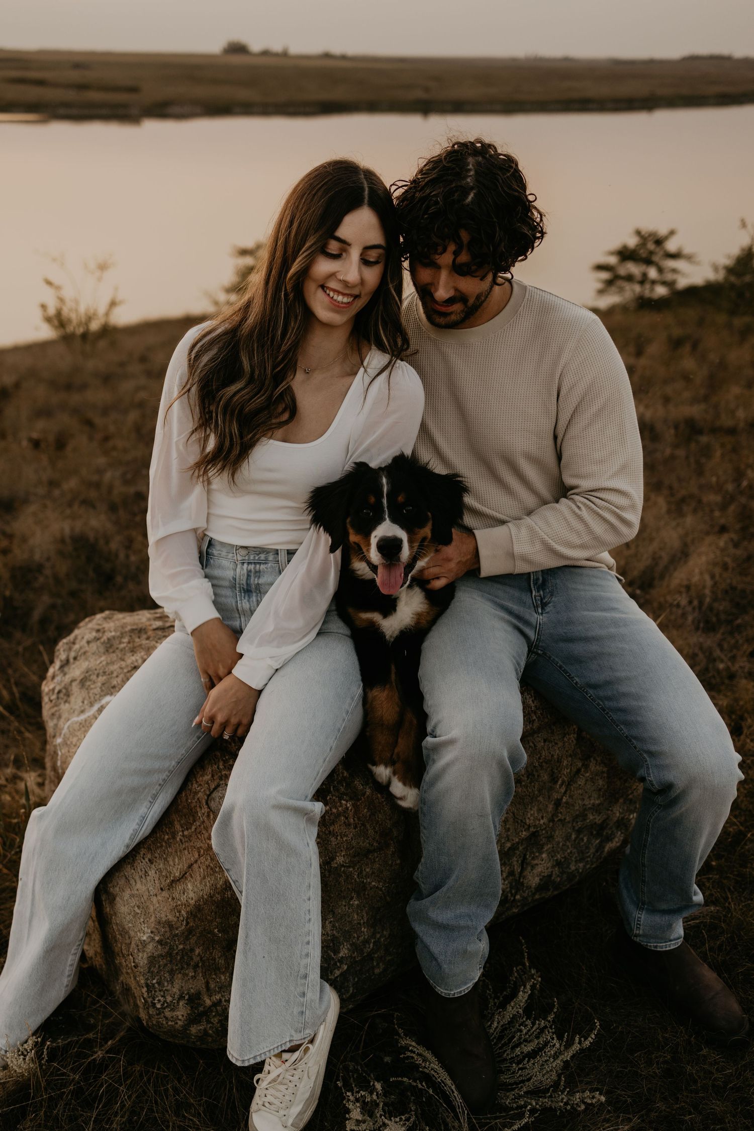 A casual couple sits on rocks with their border collie dog at sunset by a lake.