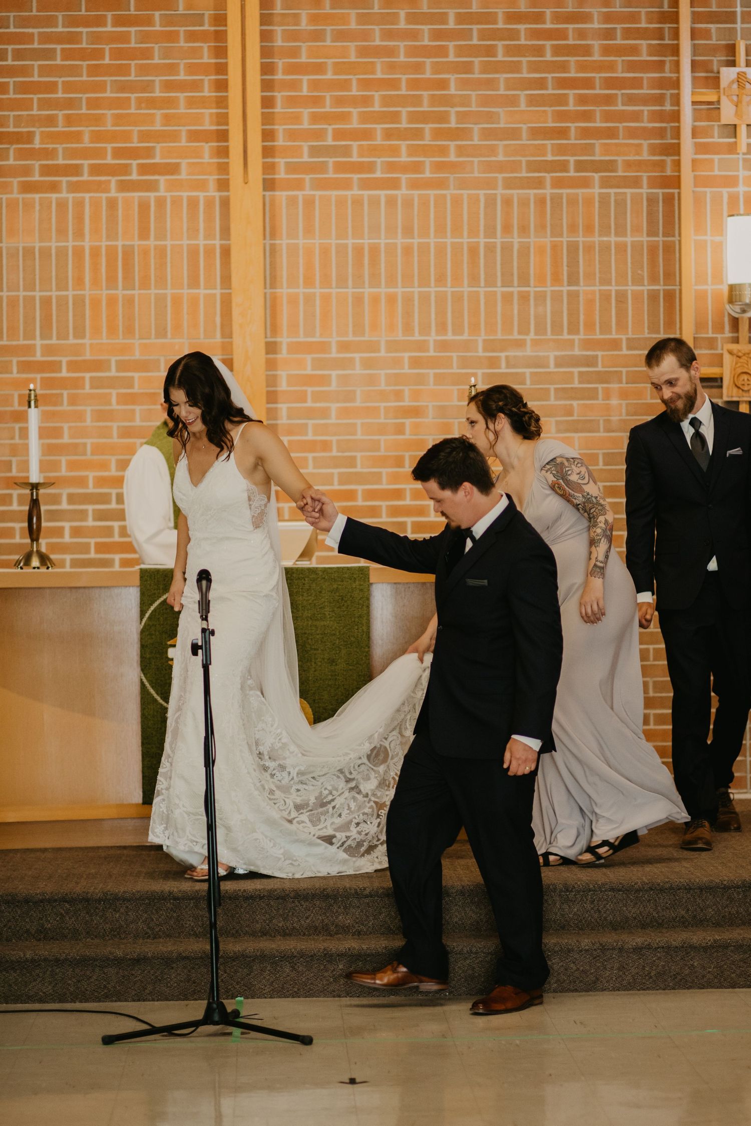 Wedding party members assist with the bride's dress train near a microphone stand.