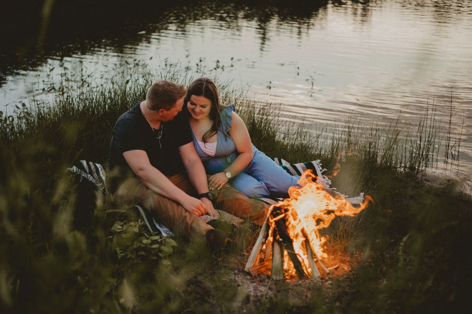 A cozy campfire scene with a couple sitting by the water at dusk.