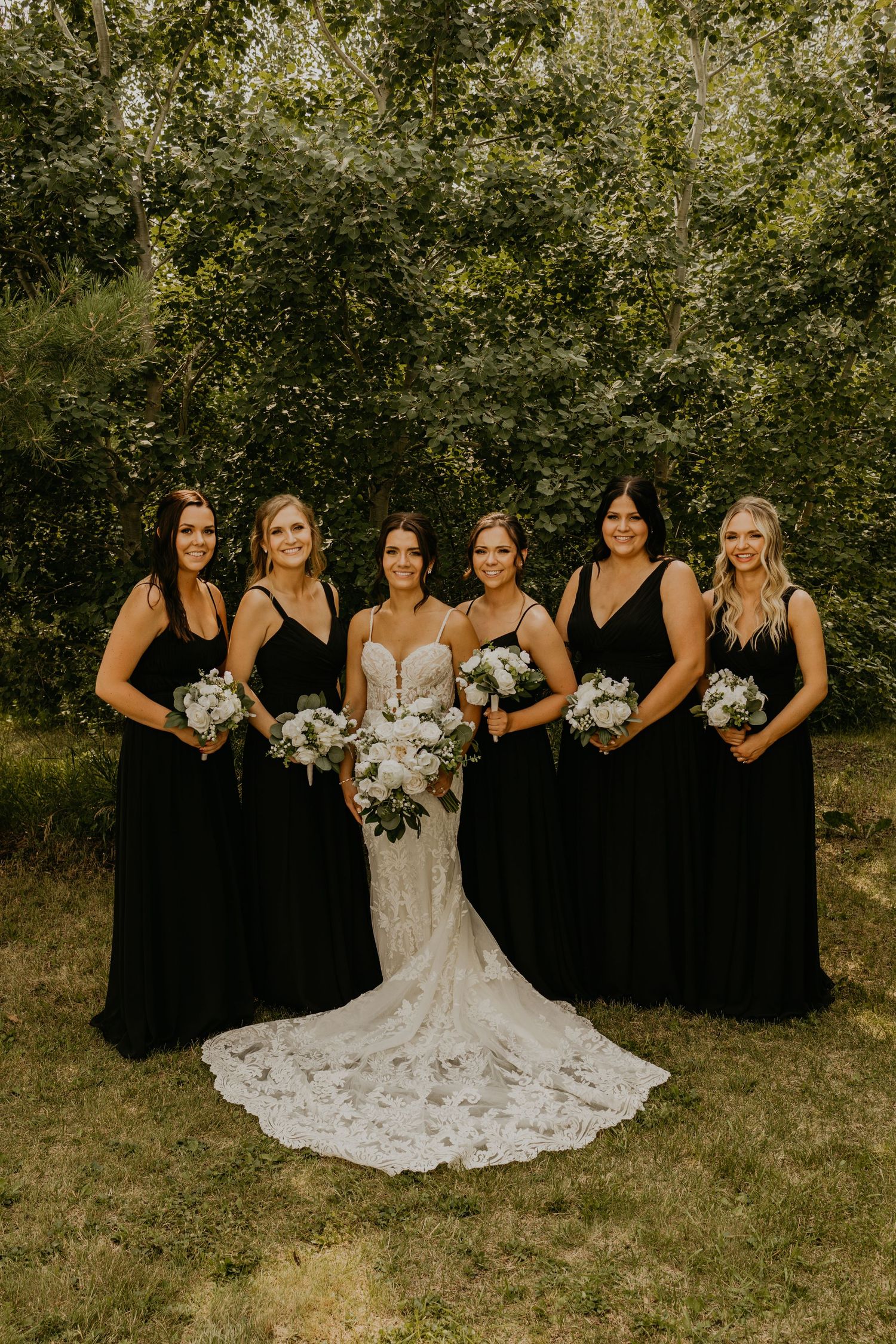 Wedding party poses outdoors in black bridesmaid dresses and white lace gown against natural greenery backdrop.