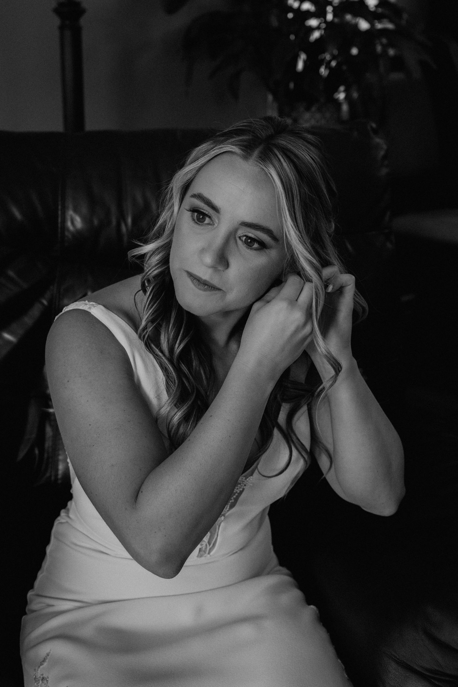 Bride in white dress sits on leather couch during wedding preparation in black and white photography.