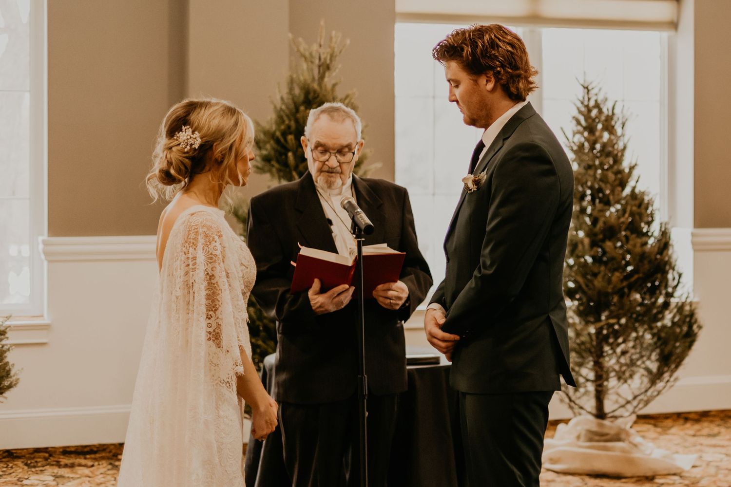 Wedding ceremony in progress with officiant and decorated Christmas trees in background.