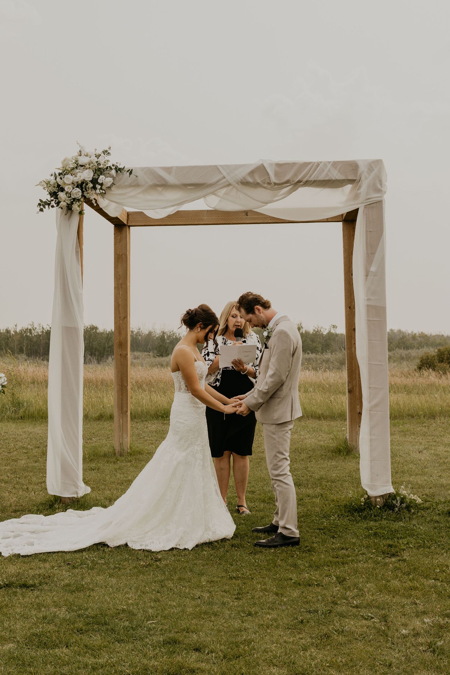 Wedding ceremony moment under a wooden arch with white draping and floral decor.