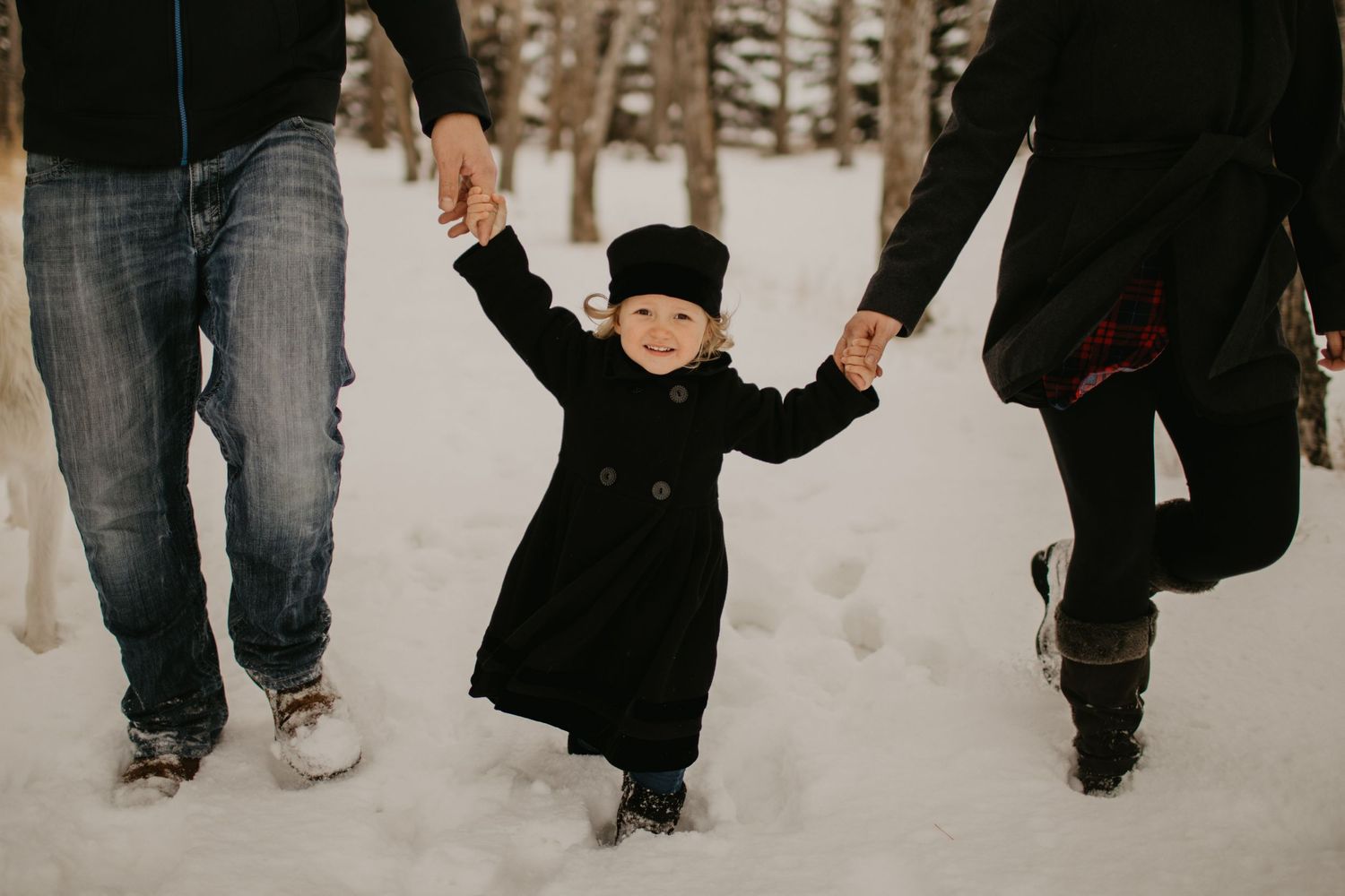 A small child in winter clothes walks through snow while holding hands with adults.