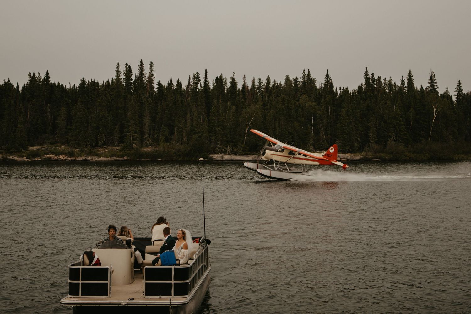 Red floatplane taking off from lake while people watch from pontoon boat.