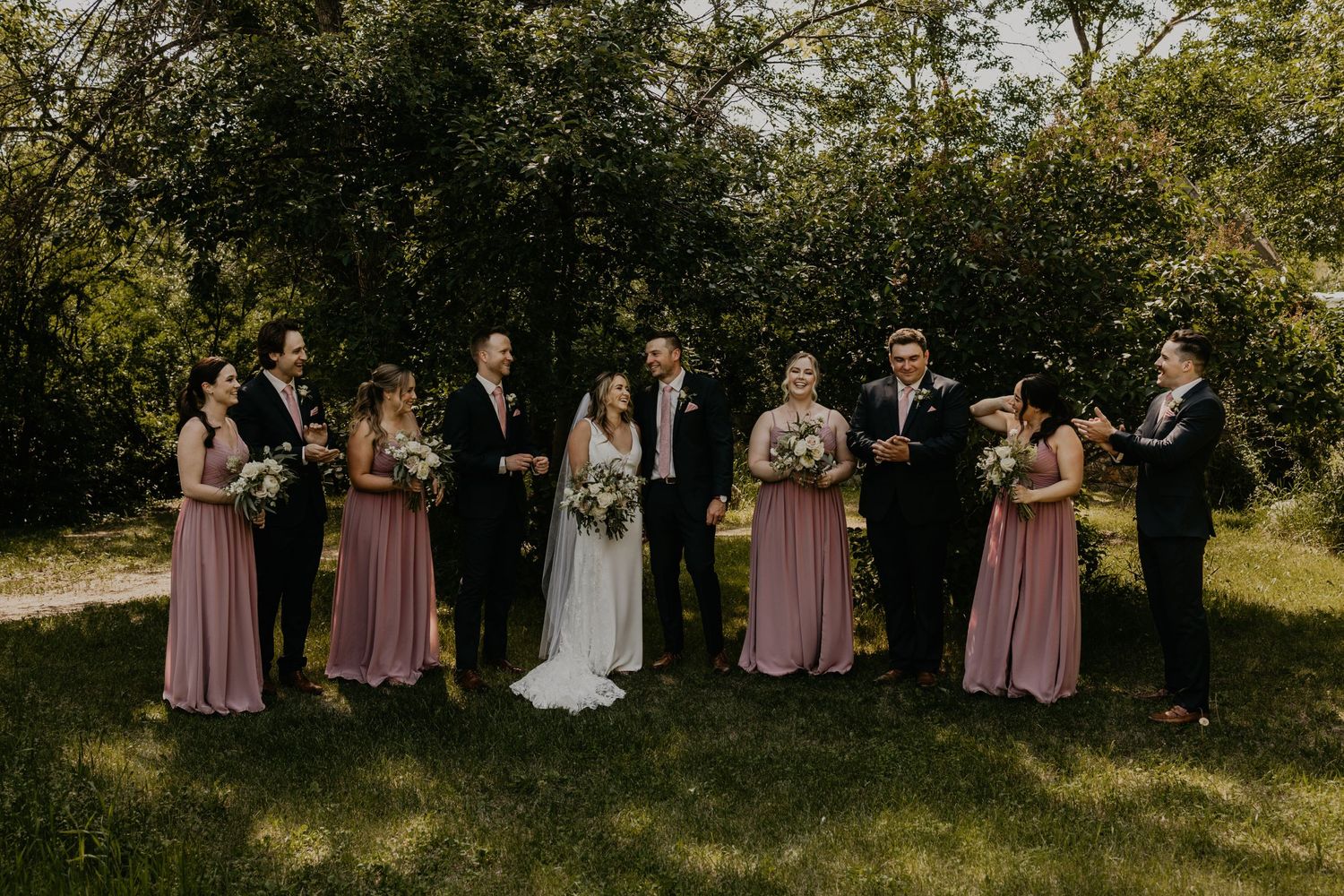 Wedding party poses on grassy lawn with bridesmaids in dusty rose dresses.