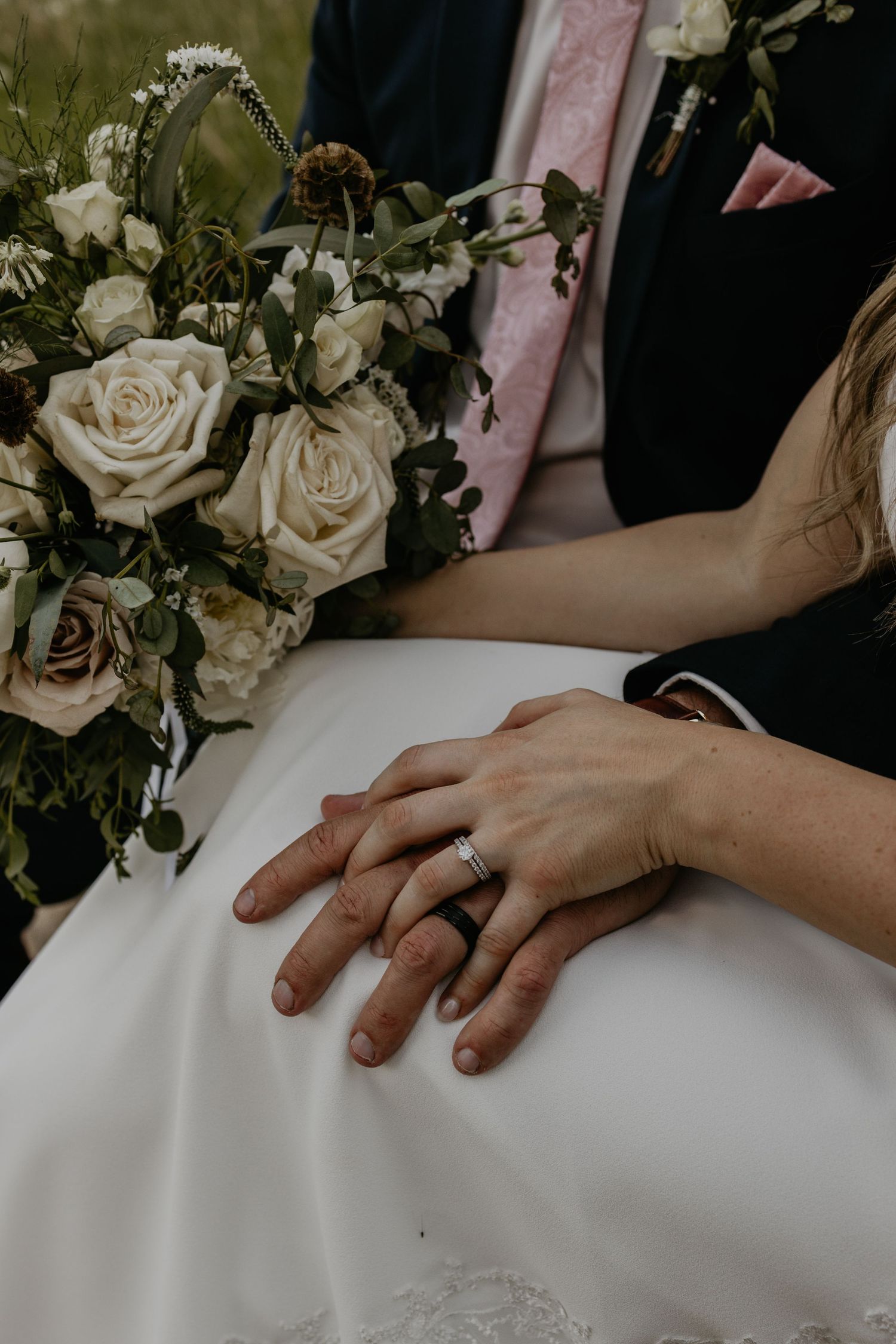 A close-up of wedding rings and hands with a white rose bouquet in the background.