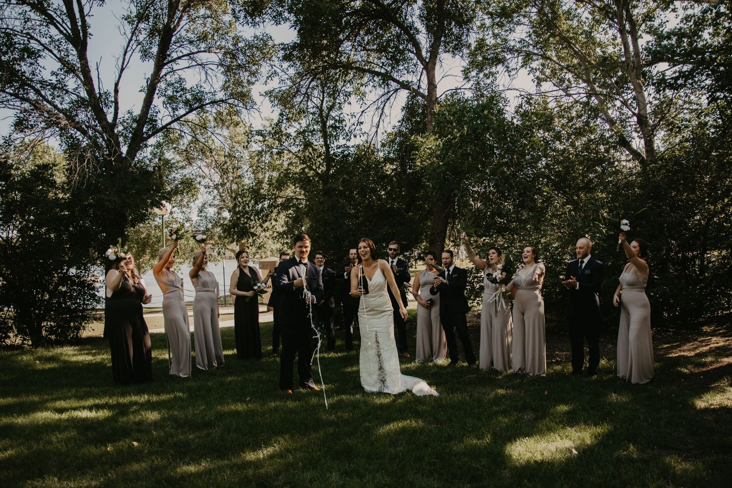 Wedding party stands together during outdoor ceremony with sunlight filtering through trees.