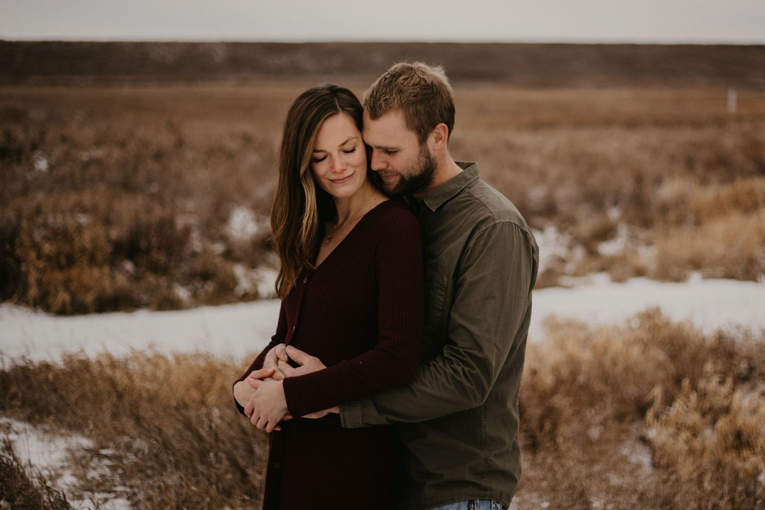 A couple embraces tenderly in a wintry field with patches of snow.
