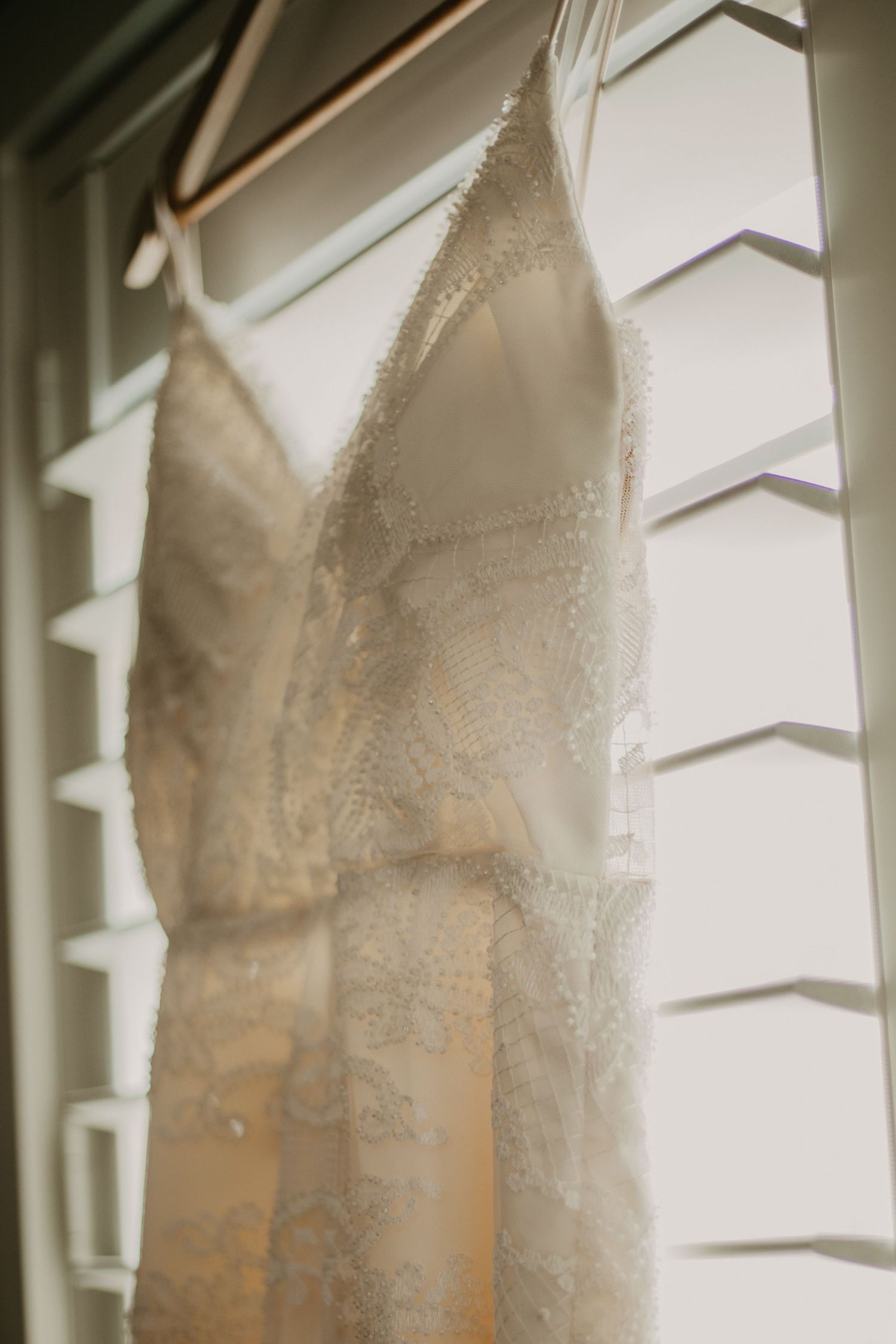 A delicate lace wedding dress hangs by white shuttered windows in natural light.