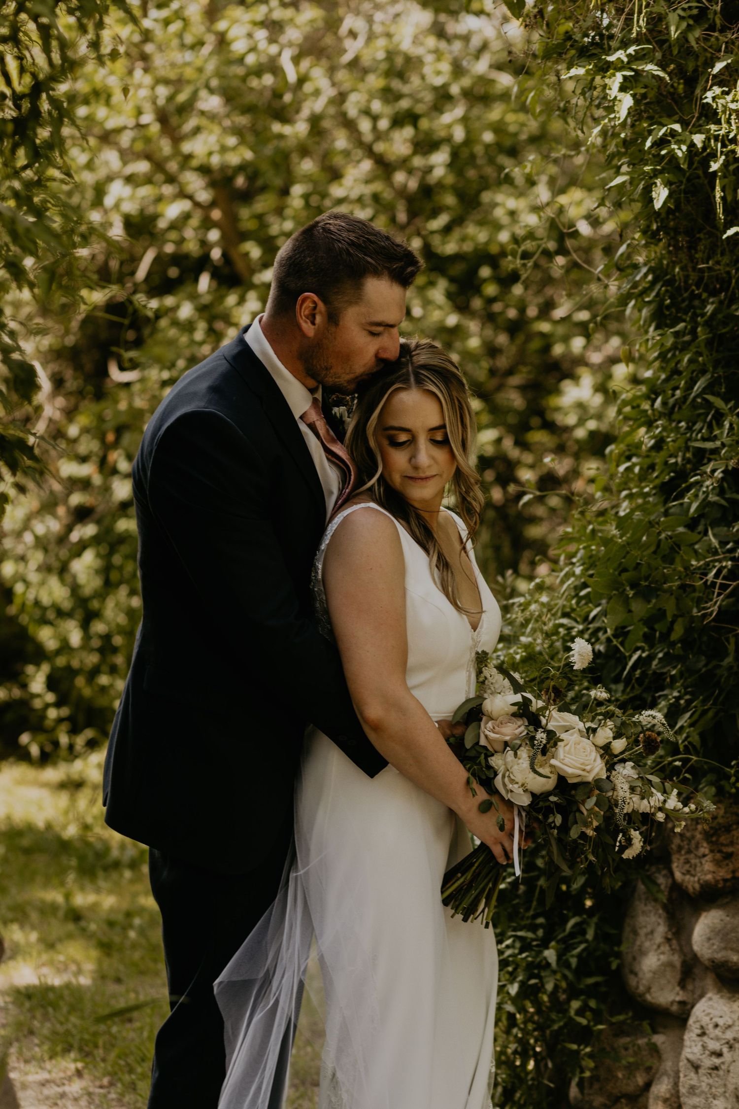 Intimate wedding moment captured against stone wall with natural greenery.