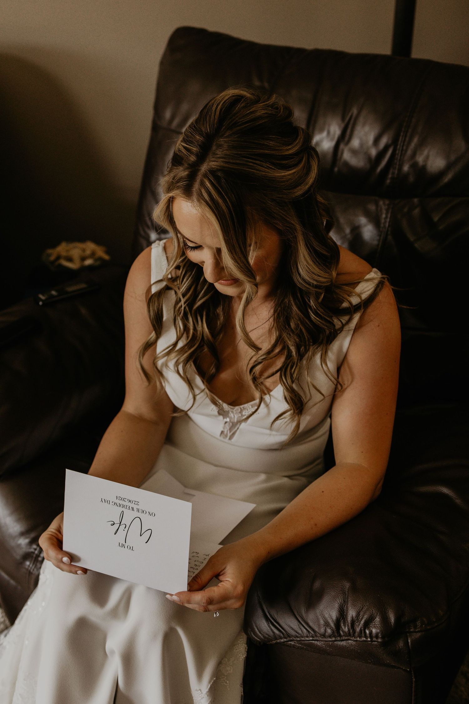 Bride in white dress reads letter while sitting on leather couch during wedding preparations.