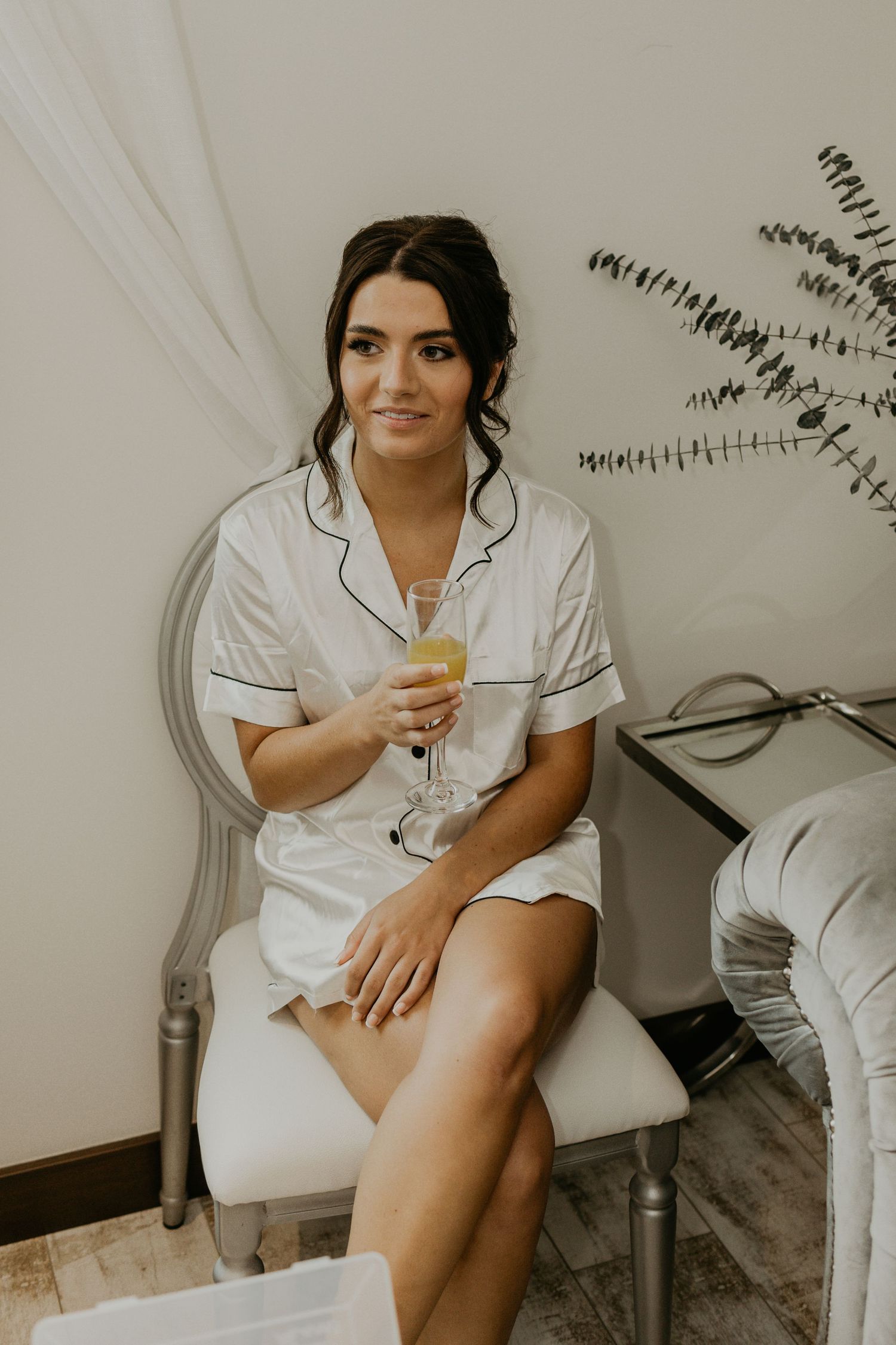 A person in white silk pajamas holds a champagne glass while getting ready.