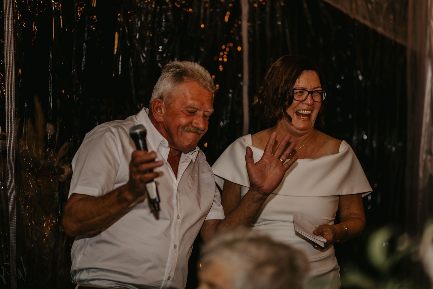 Wedding guests sharing joyful laughter and speeches during an evening reception celebration.