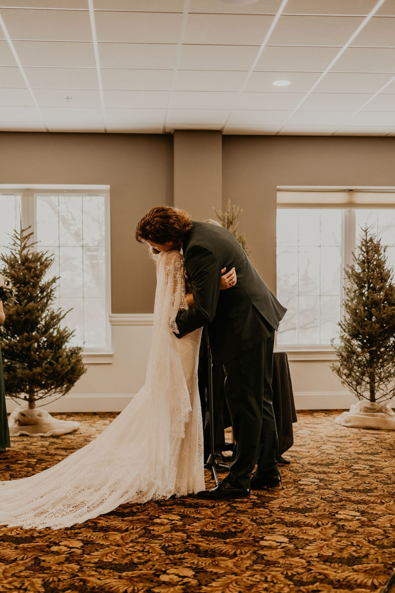 A tender moment between newlyweds with elegant Christmas trees in the background during an indoor winter wedding ceremony.