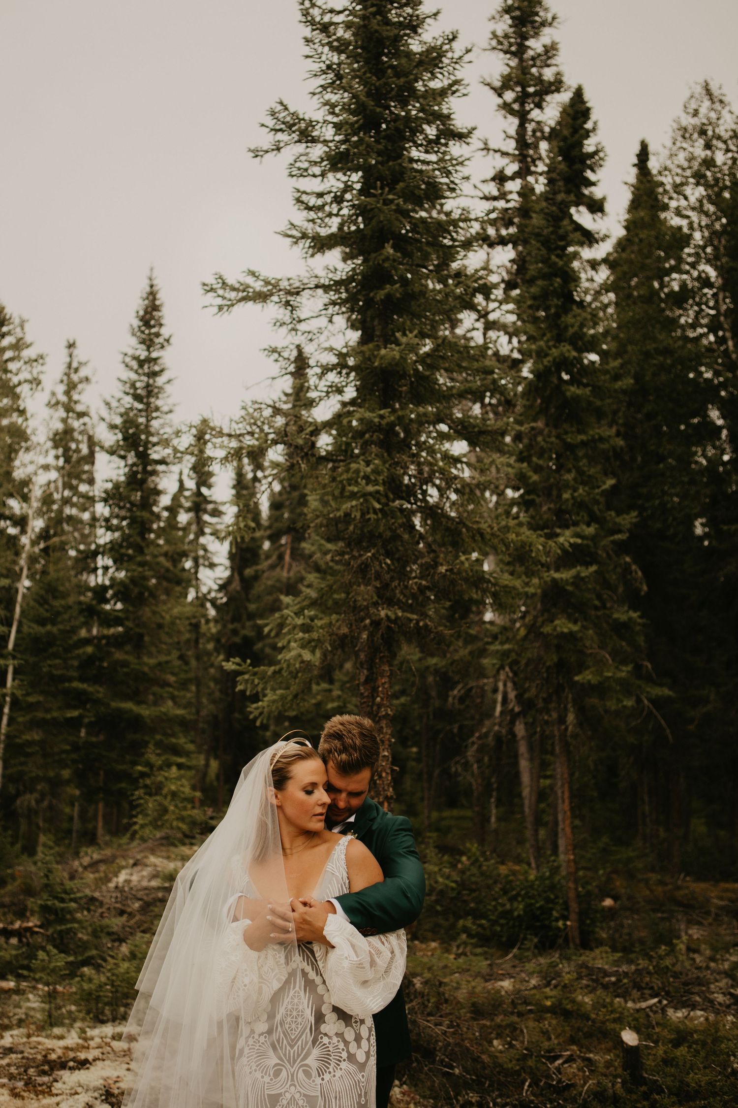 Romantic forest wedding portrait with couple embracing among tall pine trees.