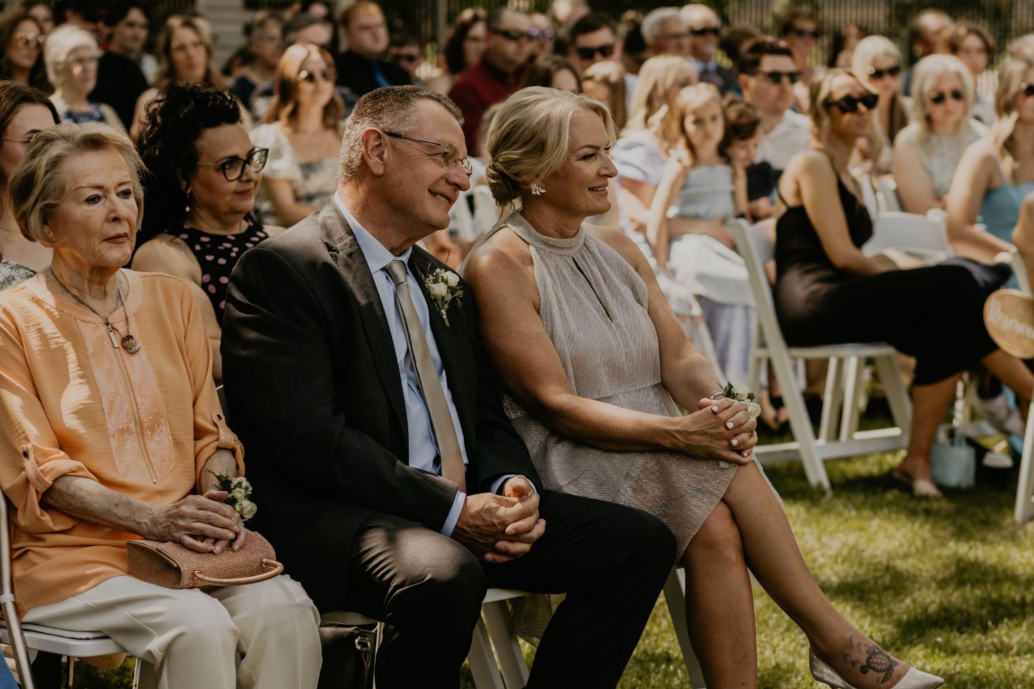 Guests watching an outdoor summer wedding ceremony while seated on white chairs.